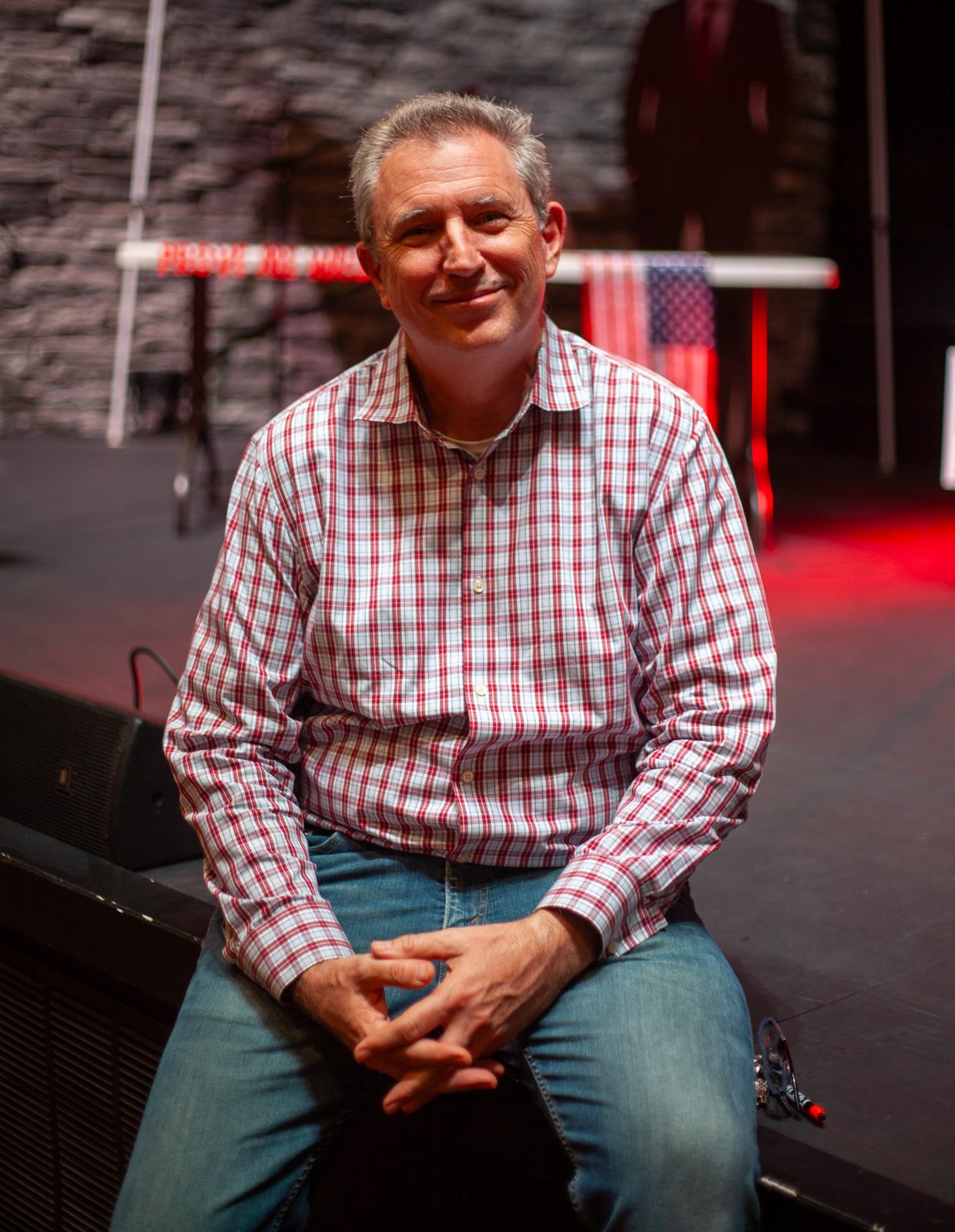 Dr. Chris Firestone seated at the edge of a stage, smiling